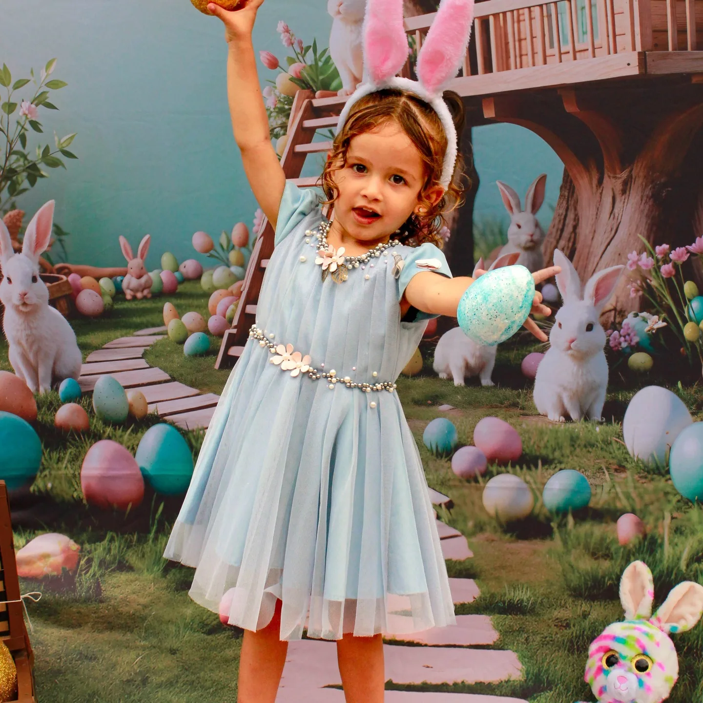 Menina de vestido azul segura um ovo colorido diante de uma decoração de festa com coelhos.