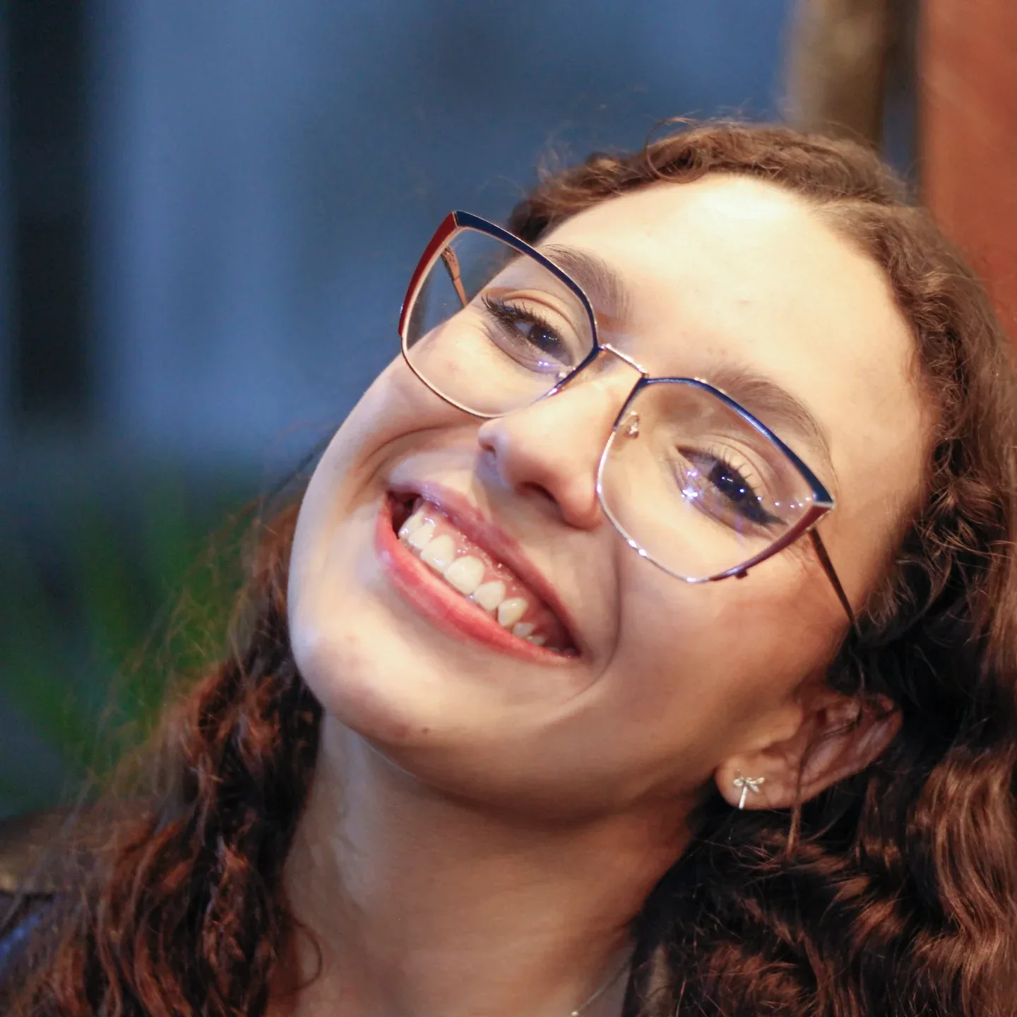 Mulher de óculos sorri diante de uma vegetação desfocada.