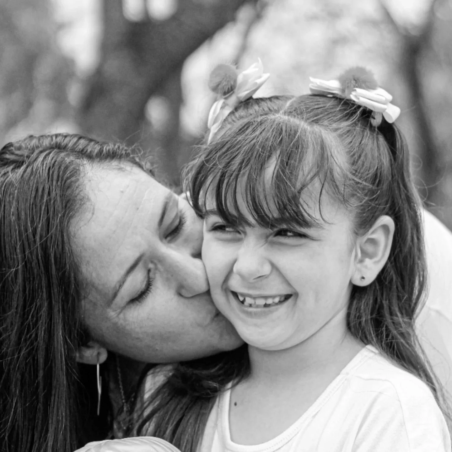 Mulher beija o rosto de uma menina sorridente, em fotografia em preto e branco.