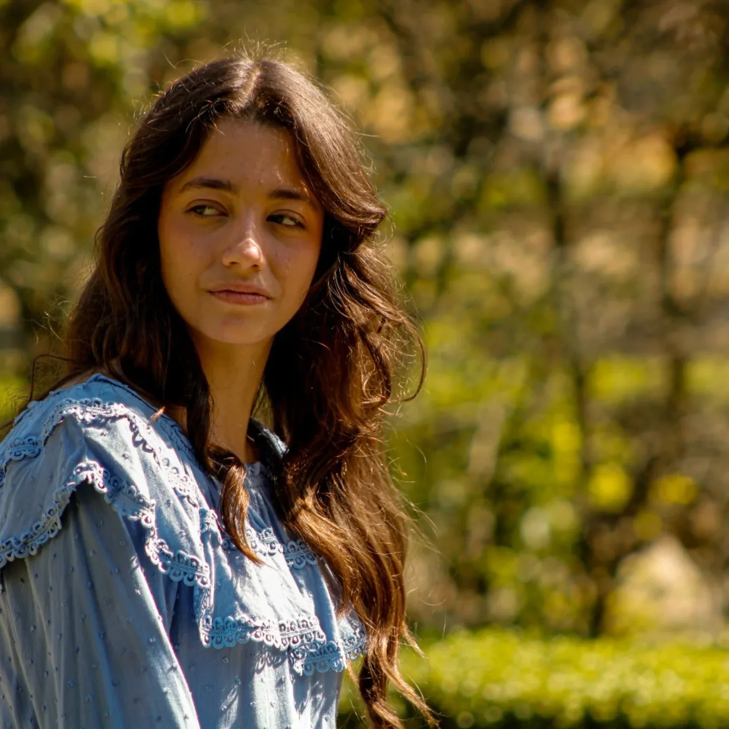 Jovem de blusa azul posa em um jardim com luz natural.