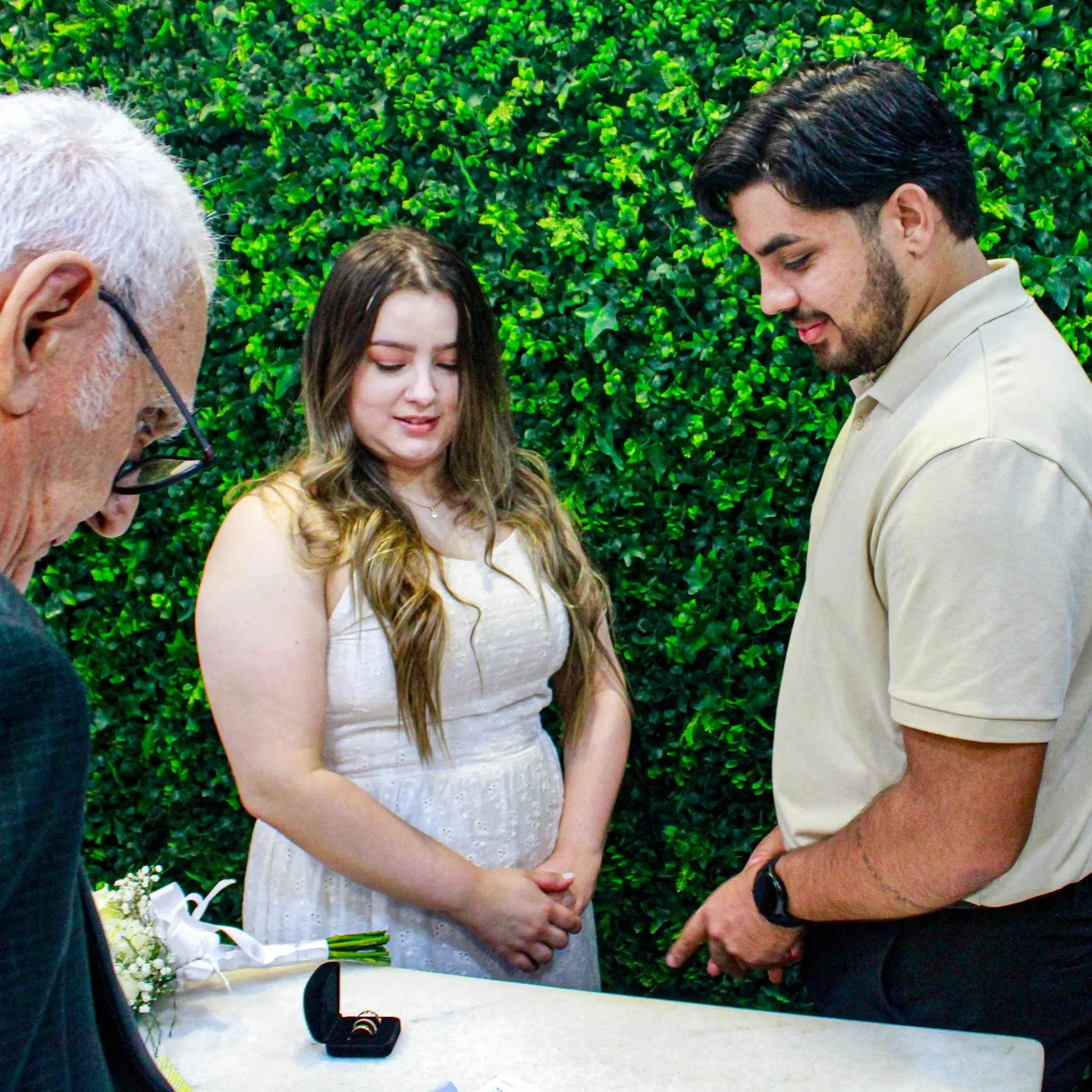 Casal acompanha uma cerimônia de casamento civil diante de uma parede de folhagens.