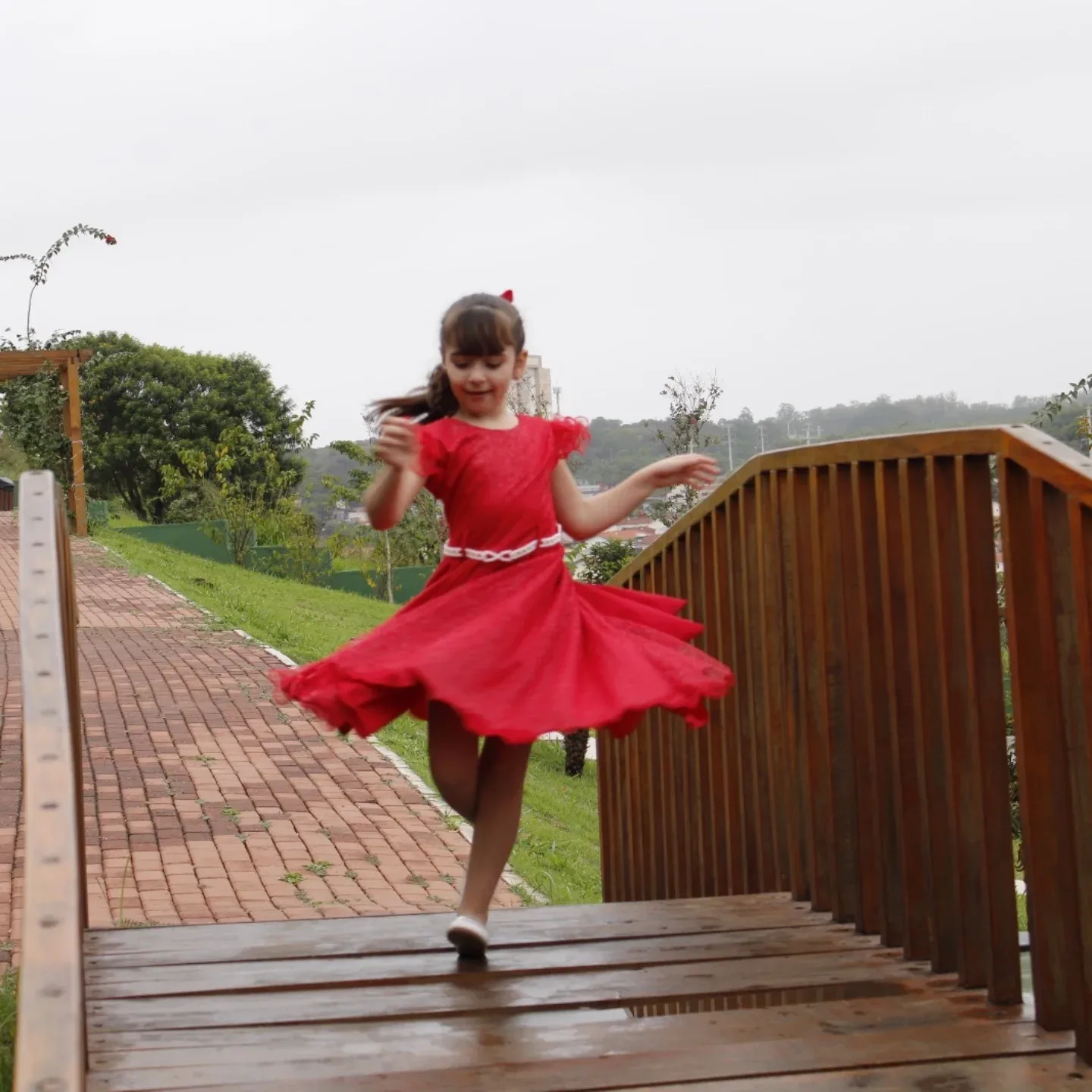 Menina de vestido vermelho gira sobre uma ponte de madeira em um jardim.