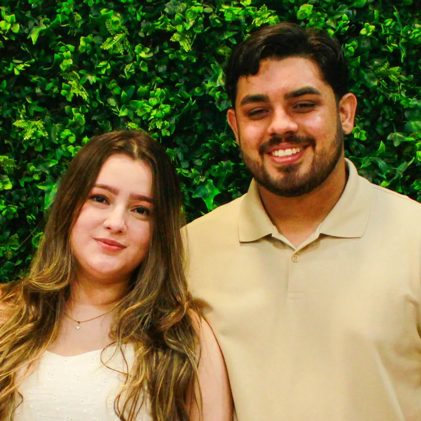 Casal sorri diante de uma parede de folhagens, com buquê em primeiro plano.