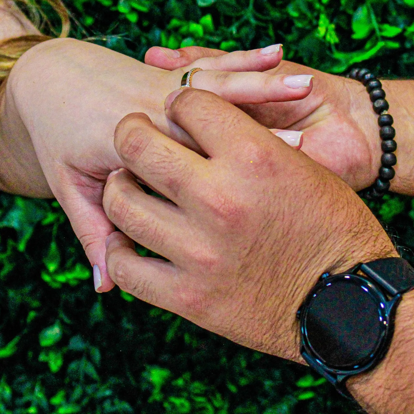 Mãos de um casal se apoiam juntas, com alianças e folhagens ao fundo.