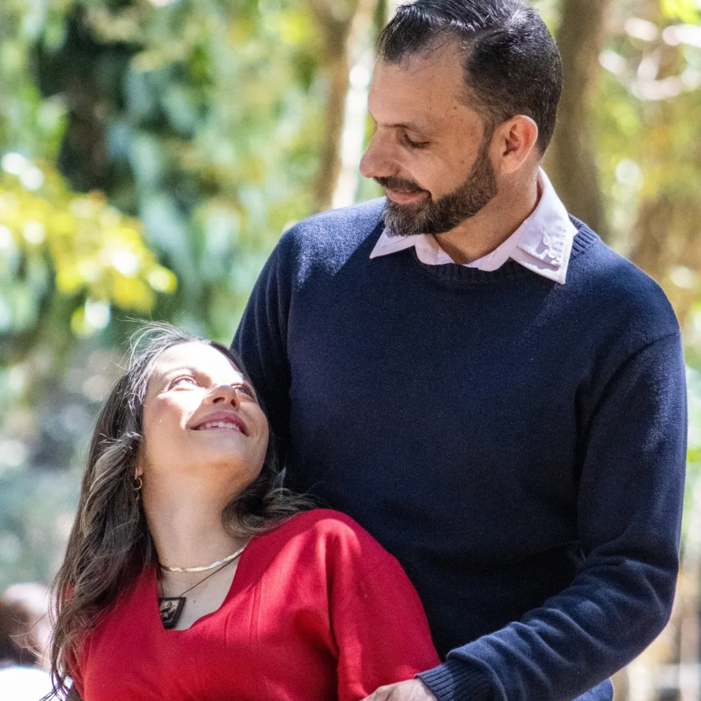 Mulher de vestido vermelho ri ao lado de um homem de blusa azul, em um jardim.
